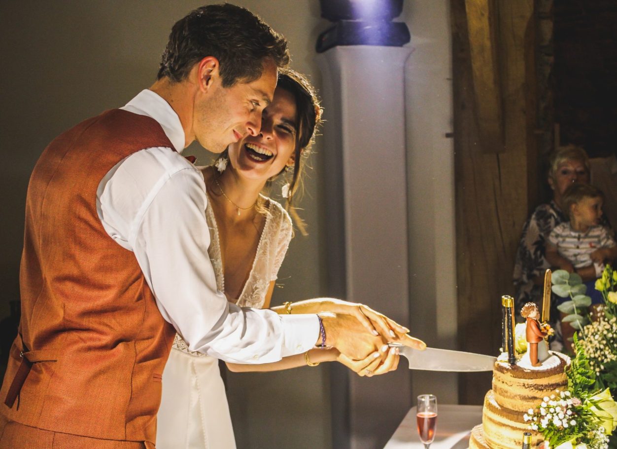 Photographe pour mariage Un couple souriant coupe un gâteau de mariage au milieu d'une célébration.